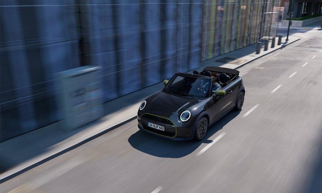 Siyah renkteki, üstü açık Mini Cooper Paul Smith Edition cabrio otomobil, direksiyon başındaki sürücüsüyle birlikte hareketli bir şehir yolunda ilerlerken, aracın modern ve dinamik tasarımı, çevresindeki hızla akıp giden yapıların hafif bulanıklığıyla vurgulanıyor.