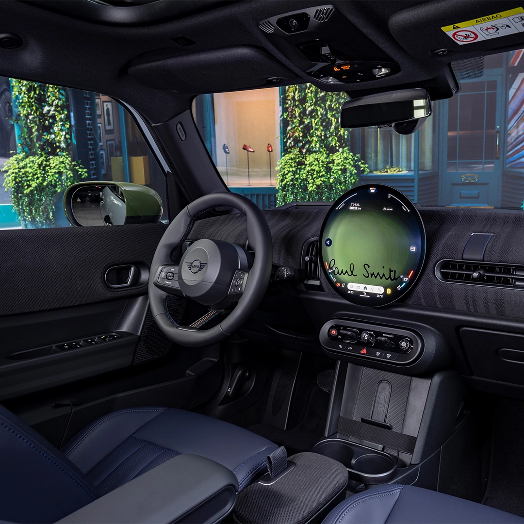 Bu MINI Cooper Paul Smith Edition iç mekan görselinde, sürücü koltuğundan görülen ahşap desenli koyu renk gösterge panelinde, ortadaki büyük dairesel ekranda Paul Smith imzası belirgin bir şekilde yer alırken, lacivert deri koltuklar ve spor direksiyon simidi modern ve şık bir atmosfer sunuyor.