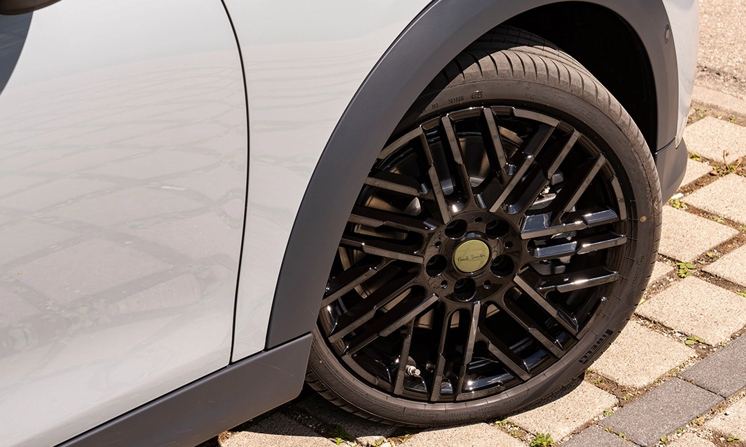Mini Cooper Paul Smith Edition aracının jantının yakın çekim görüntüsü, parlak siyah çok kollu tasarımı, ortasındaki Paul Smith logolu altın rengi kapak ve Pirelli lastiği ile aracın benzersiz estetiğini vurguluyor.