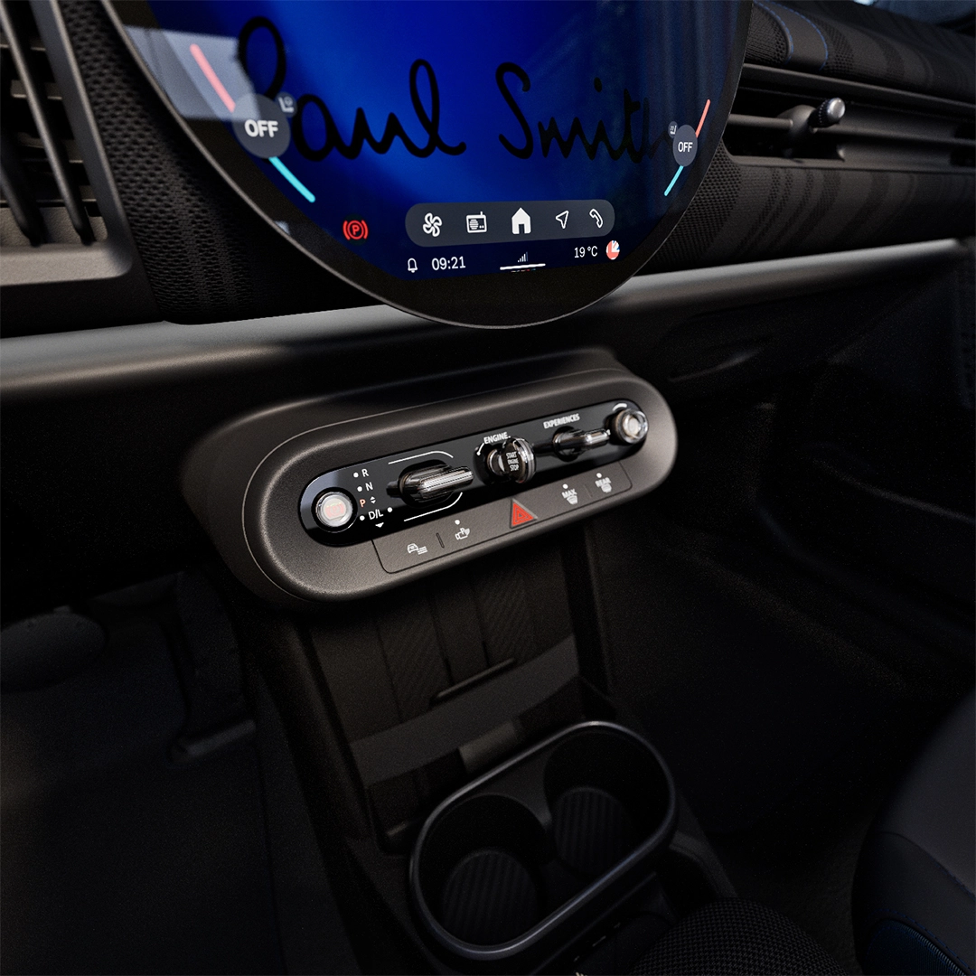 Mini Cooper Paul Smith Edition aracının iç mekanını gösteren bu detaylı çekimde, üzerinde Paul Smith imzasının bulunduğu yuvarlak dijital multimedya ekranı ile vites seçici, motor çalıştırma düğmesi ve bardak tutucuların yer aldığı modern orta konsol net bir şekilde görülüyor.