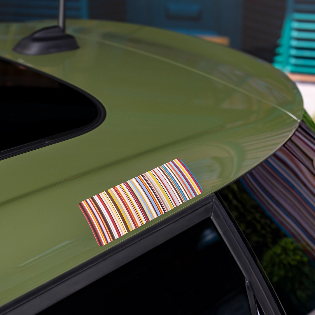 Mini Cooper Paul Smith Edition'ın zeytin yeşili tavanında, dar ve çok renkli dikey çizgilerden oluşan, canlı bir dikdörtgen çıkartma dikkat çekiyor; hemen yanında ise koyu renkli bir açılır tavan penceresi görünüyor.