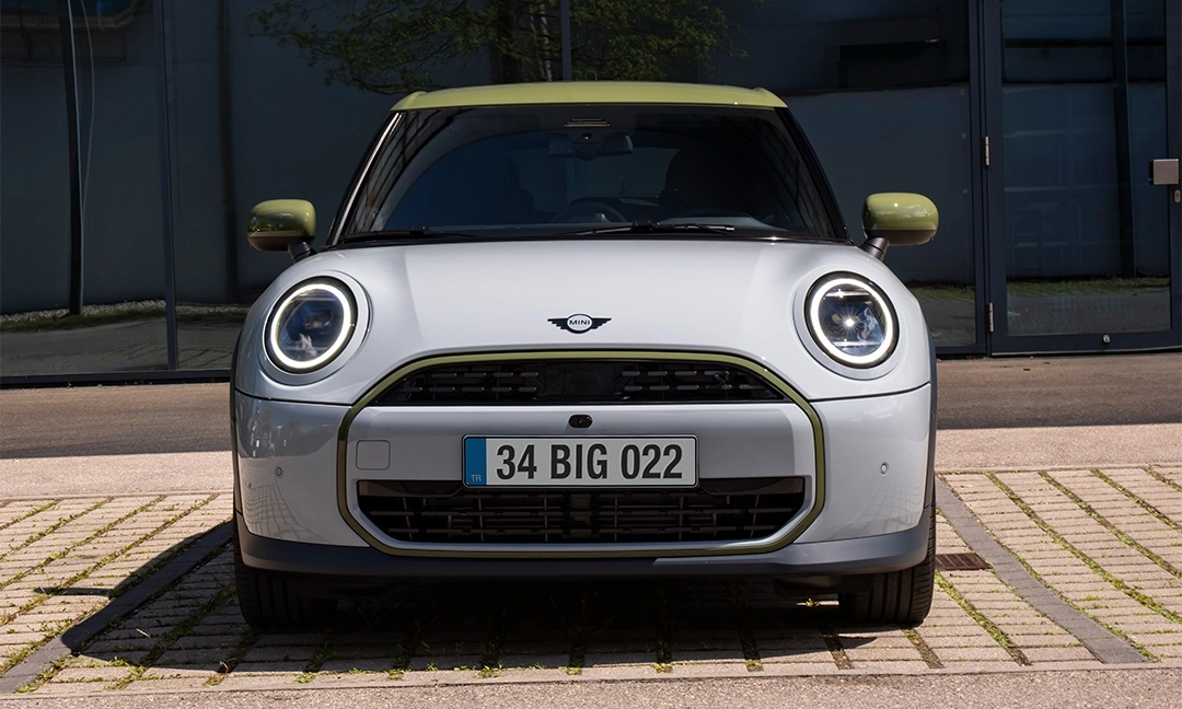 Ön planda, açık gri gövdesi, zeytin yeşili tavanı ve yan aynalarıyla dikkat çeken, modern yuvarlak LED farlara sahip özel üretim Mini Cooper Paul Smith Edition modeli, taş döşeli bir zeminde park edilmiş halde net bir şekilde görünmekte olup, arka planda yansımalı camları olan şık bir bina yer alıyor.