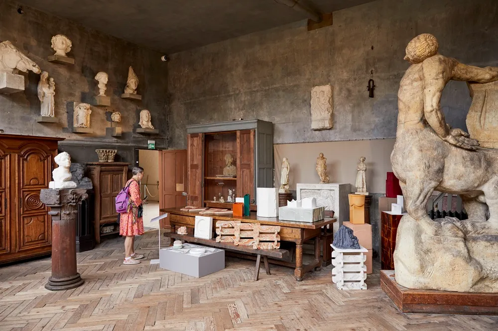 Bourdelle Müzesi'nin parke zeminli, koyu renkli duvarlara sahip tarihi bir salonunda, klasik heykellerin ve büyük sentor heykelinin ortasında Paris Tasarım Haftası için sergilenen modern tasarım objelerini dikkatle inceleyen bir ziyaretçi.