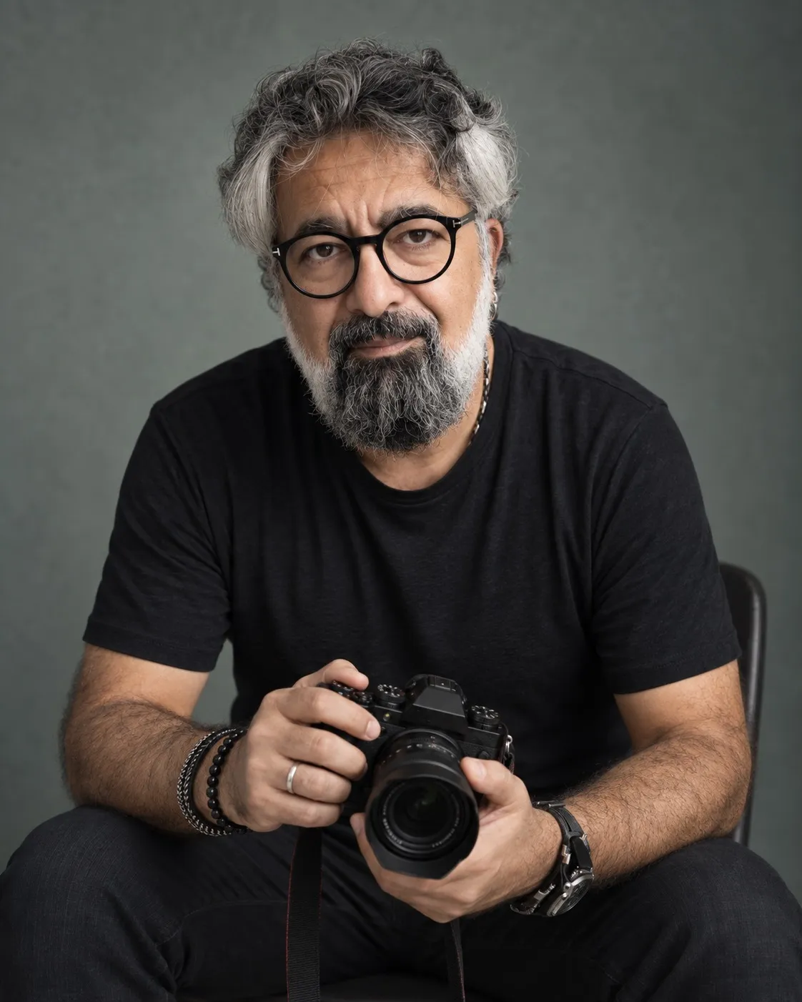 Gözlüklü, kırlaşmış saçlı ve sakallı ünlü fotoğrafçı Mustafa Seven, koyu gri bir stüdyo fonunun önünde siyah tişörtüyle otururken sol bileğindeki bileklikler ve sağ bileğindeki saatiyle dikkat çekiyor, elinde tuttuğu aynasız fotoğraf makinesiyle doğrudan izleyiciye bakıyor.