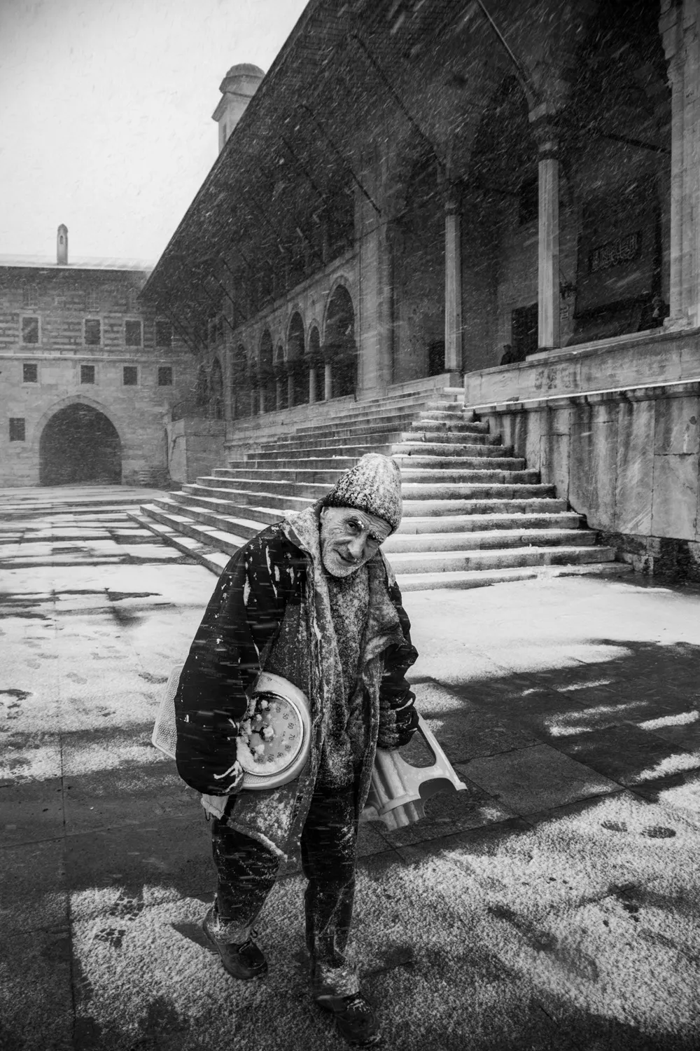 Yoğun kar yağışı altında, kalın giysileri ve örgülü beresi karlar içinde kalmış, yüzünde samimi bir gülümsemeyle kameraya bakan yaşlı bir adam, sol kolunun altında bir tartı ve sağ elinde katlanır bir tabure taşıyarak karlı tarihi bir cami avlusunda yürüyor.