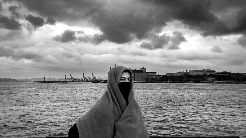 Kapalı ve bulutlu bir gökyüzünün altında, siyah beyaz fotoğrafta, yüzünü bir şalla sıkıca sarmış, sadece gözleri görünen bir kişi dalgalı denizin önünde dururken, uzaklarda liman vinçleri ve şehir binaları belirmekte.