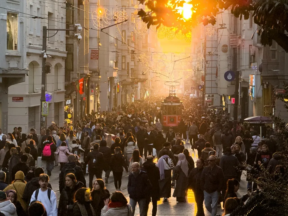 İstanbul'un ikonik bir caddesinde, gün batımının parlak altın rengi ışıkları altında ilerleyen tarihi kırmızı tramvay ve sağlı sollu sıralanmış mağazaların önünde yürüyen, cıvıl cıvıl kalabalıklar.