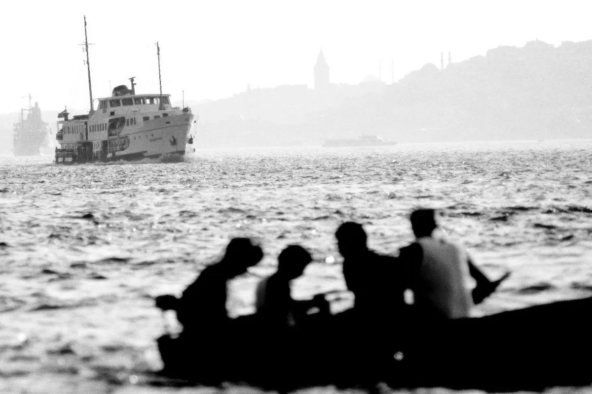 Mustafa Seven'in NASİP Sergisi'nden bir karede, sisli İstanbul Boğazı'nda ilerleyen bir vapurun ve uzaklardaki Galata Kulesi'nin önünde, bir teknede oturan silüet halindeki insanlar, şehrin gündelik akışına ve geçici anlara tanıklık ediyor.