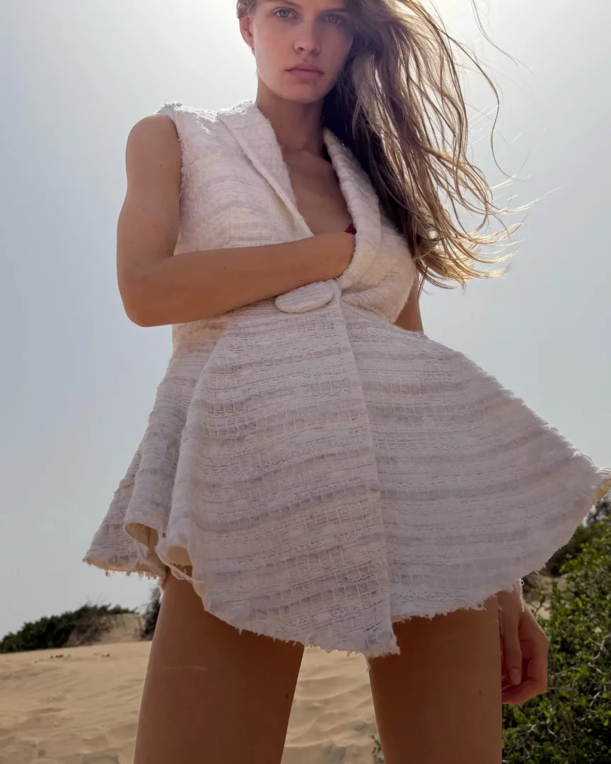 Düşük bir açıdan çekilmiş fotoğrafta, rüzgarda dalgalanan uzun saçları ve beyaz renkli, dokulu, kolsuz, kloş etekli kısa ceket elbiseyi giyen genç model, kumlu zeminde kameraya doğrudan bakarak yeni moda tasarımını sergiliyor.