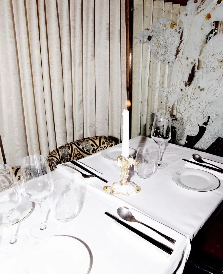 Ojii Paris restoranında beyaz masa örtüsüyle zarifçe düzenlenmiş bir yemek masası; üzerinde yanan ince bir mum, parıldayan şarap kadehleri, su bardakları ve siyah yemek çubukları ile metal kaşıklar şık bir atmosfer yaratıyor.