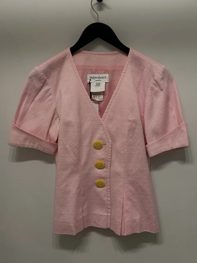Açık pembe renkli, hafif kabarık kısa kollu ve kıvrık manşetli, önünde üç sarı düğme bulunan, zarif desenli kumaşa sahip vintage bir Yves Saint Laurent bluzu, Paris modasının inceliğini yansıtır şekilde bir askıda sergileniyor.