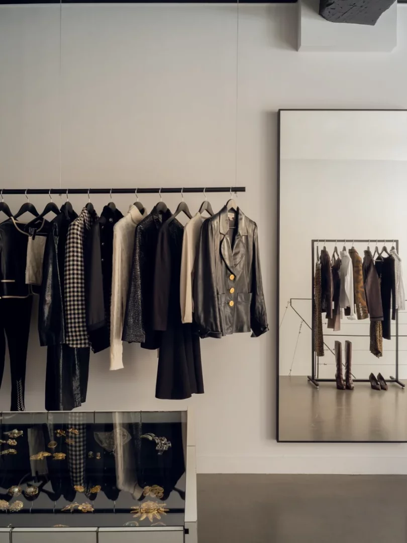 Beyaz duvarlı, modern bir butikte, tavandan sarkan siyah bir askılıkta koyu renkli şık kıyafetler, altında ise cam vitrinde çeşitli vintage takılar sergileniyor; sağda ise aynı kıyafetlerin yansıdığı büyük bir ayna ve yerde çizmeler duruyor, adeta Paris esintili bir vintage mağaza atmosferi sunuyor.