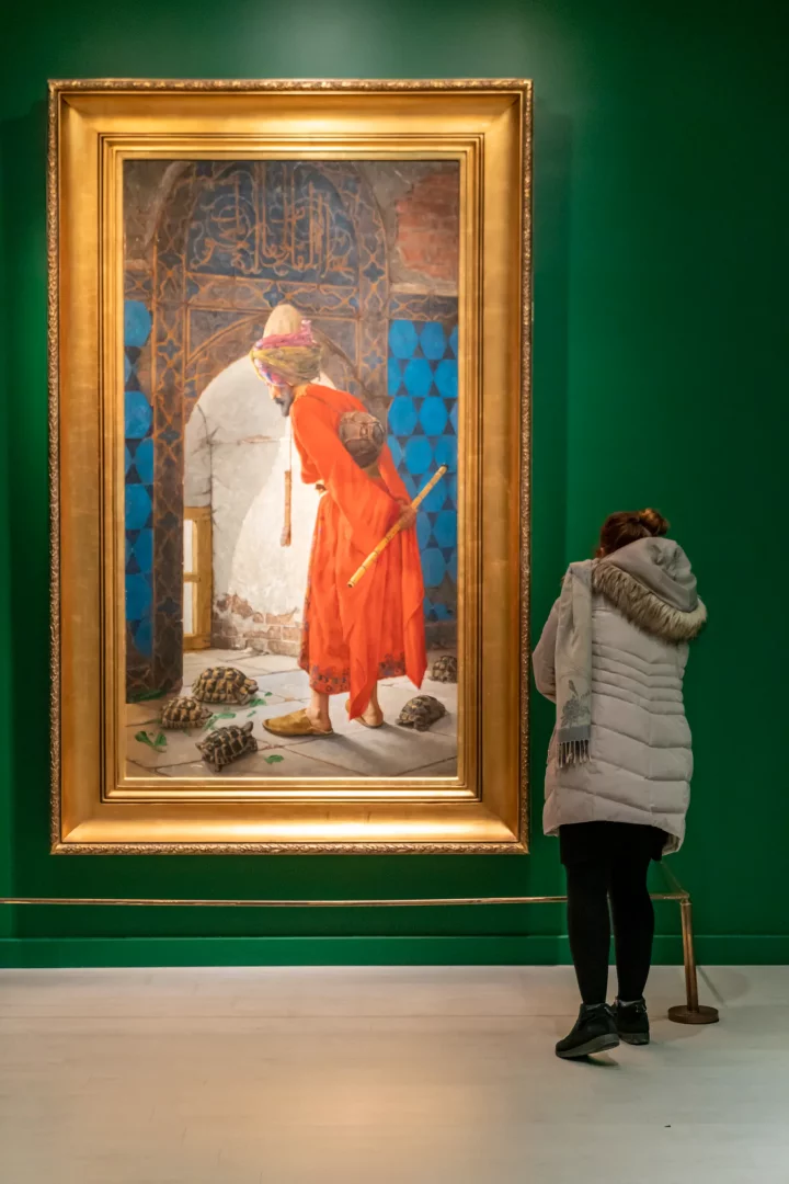 Yeşil duvarlı bir salonda, arkası dönük bir ziyaretçi, Pera Müzesi'nin Oryantalist Resim koleksiyonundan Osman Hamdi Bey'in ünlü Kaplumbağa Terbiyecisi tablosuna dikkatle bakarken, bu an Beyoğlu kültür rotasındaki zengin sanat duraklarından birini yansıtıyor.