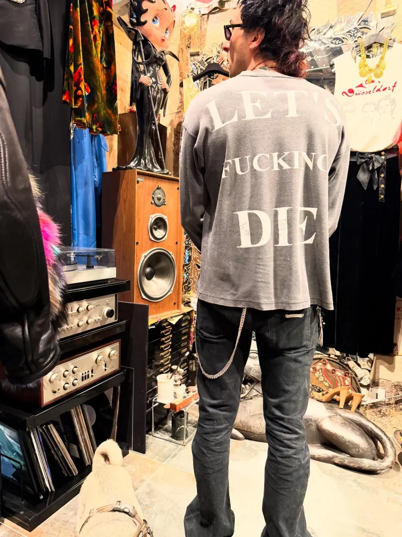 Sırtı dönük, üzerinde beyaz büyük harflerle LET'S FUCKING DIE yazan gri uzun kollu bir tişört ve zincirli koyu renk kot pantolon giyen bir kişi, içindeki dekoratif eşyaların ve kutuların dikkat çektiği, ahşap bir hoparlörün üzerinde duran siyah elbiseli Betty Boop figürü, sol alt köşede yer alan pikap ve amplifikatörlerden oluşan ses sistemi, dikey depolanmış vinil plaklar ve yere bakan küçük bir köpek ile dolu, kalabalık ve vintage bir mağaza ortamında durmaktadır.