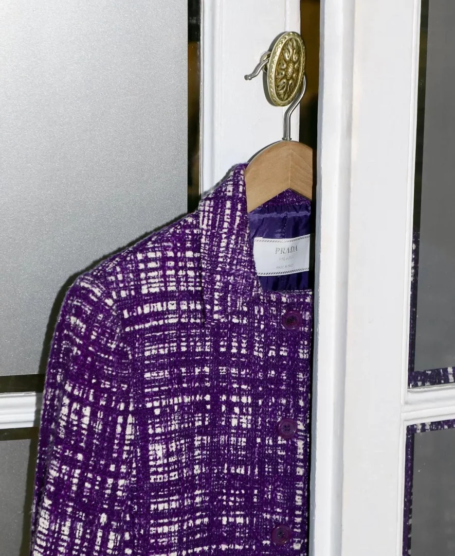 Beyaz kapı pervazındaki dekoratif pirinç askıya asılmış, mor ve beyaz renklerin iç içe geçtiği tweed kumaş dokusuna sahip Prada marka ceket, lüks moda ve zarif tasarım anlayışının özgün bir temsilini sunuyor.