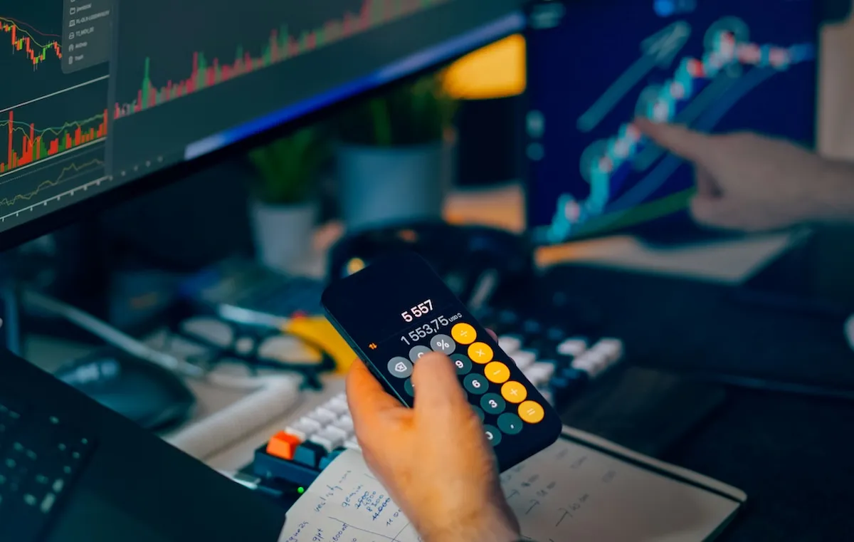 Bir yatırımcı, elindeki akıllı telefondaki hesap makinesinde 1553,75 USD gibi değerleri hesaplarken, bilgisayar ekranlarındaki candlestick grafikleri ve detaylı finansal verileri dikkatle takip ederek, portföyündeki risk değeri ve fon seçimi kararları için kapsamlı bir analiz gerçekleştiriyor.