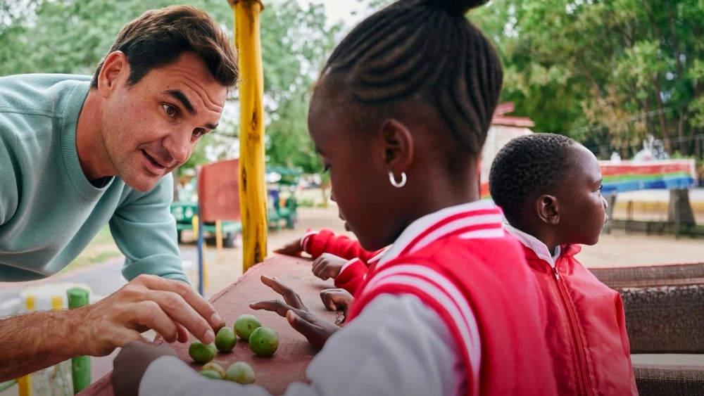 Açık mavi bir kazak giyen Roger Federer, gülümseyerek eğilmiş bir şekilde, kırmızı ve beyaz ceketli, örgülü saçlı küçük bir çocuğa yeşil taneleri gösterirken, Roger Federer Vakfı'nın çocuklara olan desteğini yansıtan bu anı arka planda bir başka çocuk izliyor.
