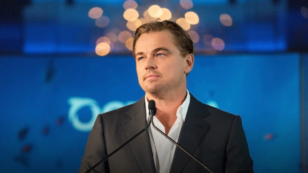Leonardo DiCaprio, koyu renk takım elbisesi ve beyaz gömleğiyle, ReWild Vakfı için düzenlenen bir bağış etkinliğinde mikrofonun önünde dururken, bulanık mavi bir arka plan ve ışık huzmeleri önünde düşünceli bir ifadeyle yukarı doğru bakıyor.