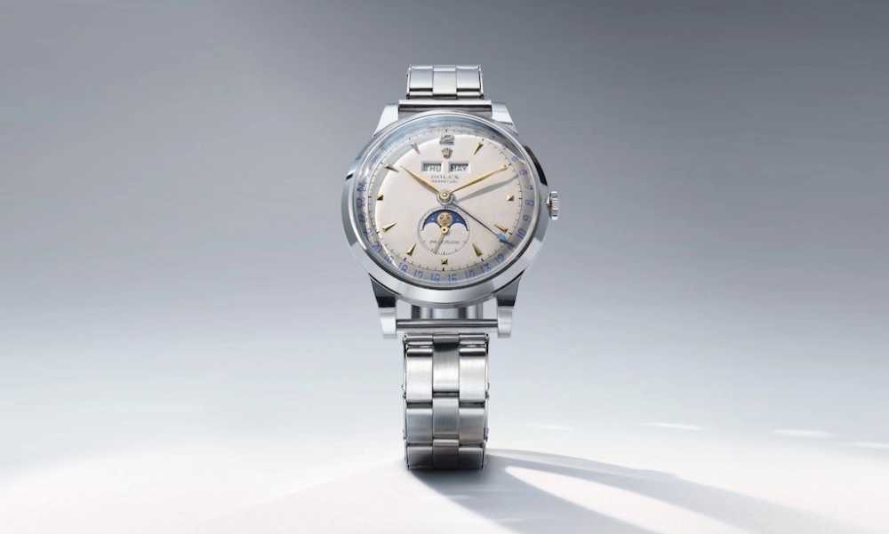 Ortalı bir açıyla beyaz bir arka plan üzerinde duran, gümüş renkli metal bileziğe sahip, kremsi kadranında ay evresi, gün ve ay göstergeleri bulunan bu ikonik 1951 model Rolex Padellone klasik vintage saat, zarif detaylarıyla göz kamaştırıyor.