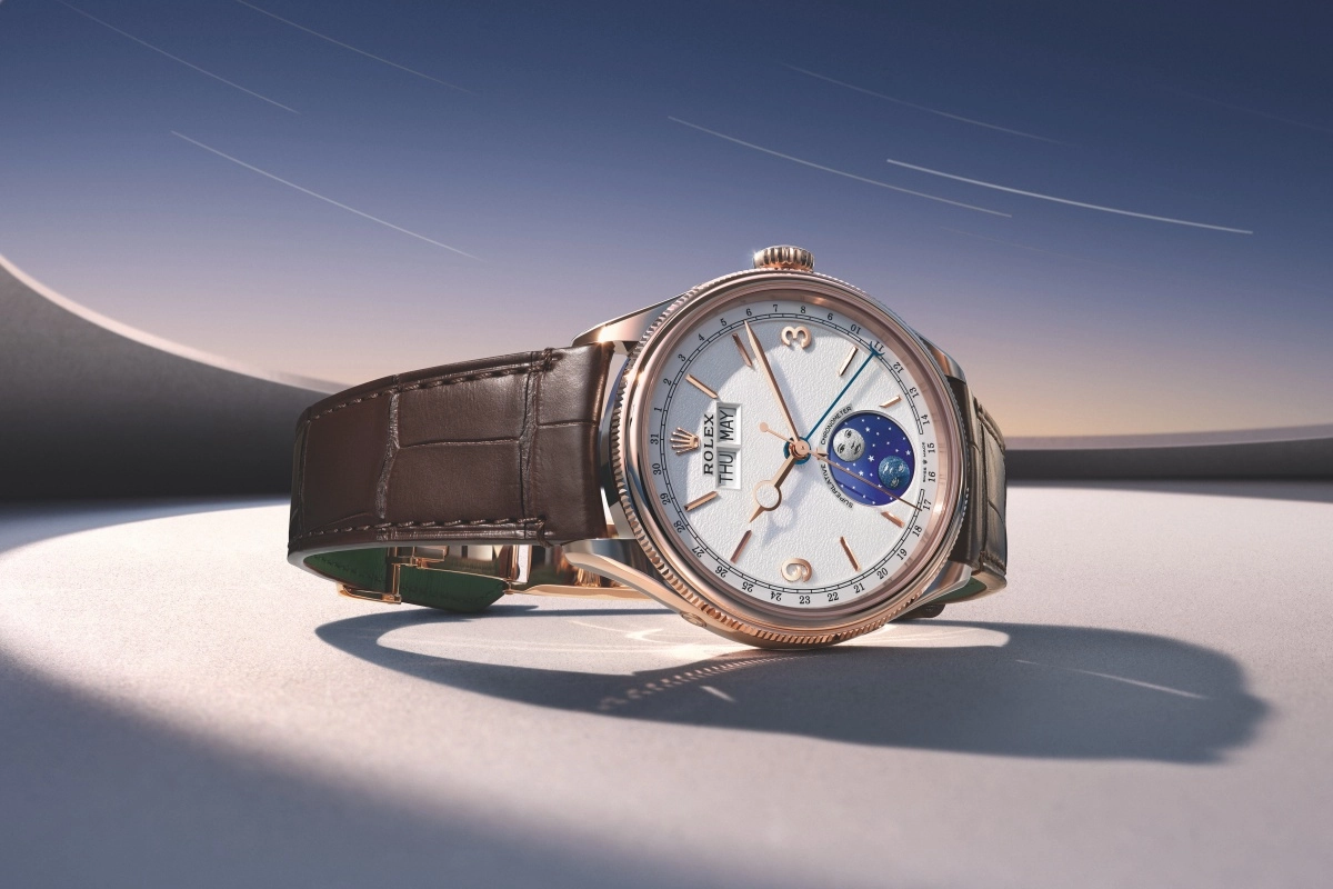 Rolex Perpetual Padellone Evrose Altın kasalı, beyaz dokulu kadranında gün (Perşembe), ay (Mayıs) ve detaylı ay fazı göstergeleri bulunan bu özel kol saati, kahverengi timsah desenli Deri Kayış ile şık bir zeminde, uzun gölgeler ve arka plandaki yıldız izleri ile soyut mimari öğelerin bulunduğu dingin bir atmosferde sergileniyor.