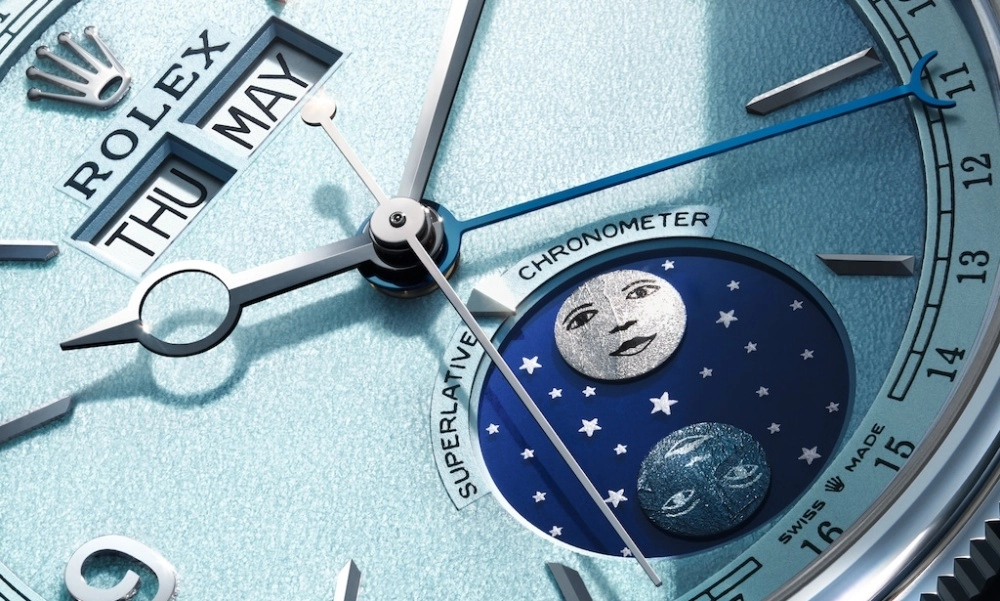 Bu yakın çekim görüntüde, Rolex Perpetual Padellone modelinin detaylı mavi kadranı ön planda olup, üst kısmında THU MAY göstergesi, merkezinde akrep ve yelkovan ile mavi ibre, alt kısmında ise karanlık gökyüzünde parlayan yıldızlar ve gümüş renkli iki ay figürü içeren zarif ay fazı göstergesi net bir şekilde görülmektedir; saatin platin kasa detayı da hafifçe seçilmektedir.