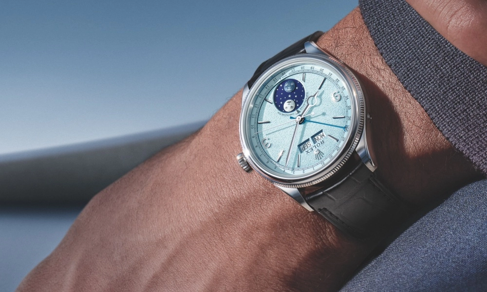 Buzlu mavi kadranı, ay fazı, gün ve ay göstergeleriyle dikkat çeken, platin kasa ve tırtıklı bezeli olan Rolex Perpetual Padellone kol saati, kroko desenli siyah deri kayışıyla bir erkeğin kolunda, hafif bulanık mavi bir arka plan önünde zarif bir şekilde konumlanmış.