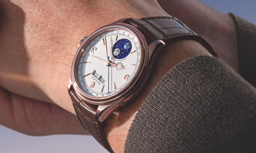 Yakın çekimde bir bilekteki, rose altın kasalı, beyaz kadrana, ay fazı ve gün-tarih göstergelerine sahip, kahverengi deri kayışlı zarif Rolex Perpetual Padellone kol saati, koyu renkli örgü bir kazağın koluyla dikkat çekici bir uyum sergiliyor.