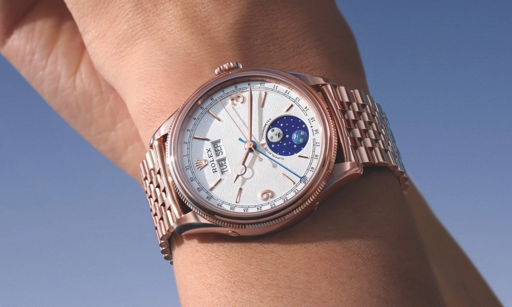Bir kişinin bileğinde, rose altın kasalı, yivli bezelli ve Jubilee bileziği bulunan, beyaz kadranında ay fazı göstergesi, gün, tarih ve mavi saniye ibresi detaylarıyla zarif Rolex Perpetual Padellone saatinin yakın çekimi.