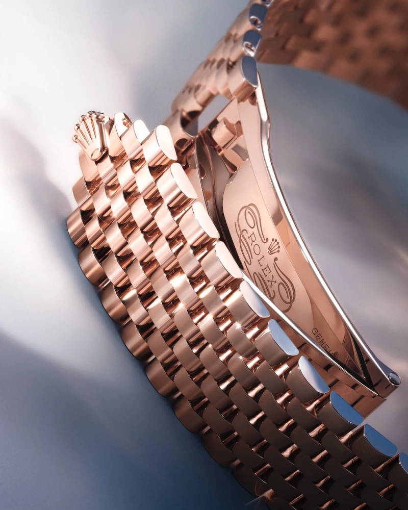 Parlak, roze altın tonlarındaki Rolex bileziğin açık kilit mekanizması ve üzerine titizlikle işlenmiş Rolex logosu ile Geneve yazısı dikkat çekiyor; yuvarlak hatlı bağlantılar ve tokadaki yansımalar, bu lüks aksesuarın detay işçiliğini vurguluyor.