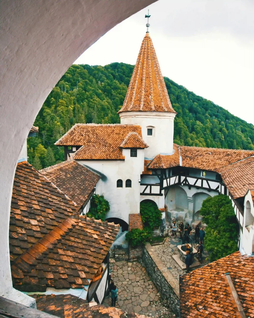 Yüksek bir noktadan çekilen bu geniş açılı fotoğrafta, Romanya'nın yemyeşil dağlarla çevrili, portakal renkli kiremit çatılı kulelere ve beyaz duvarlara sahip tarihi Bran Şatosu'nun kalabalık taş avlusu ve çevresi gözüküyor.