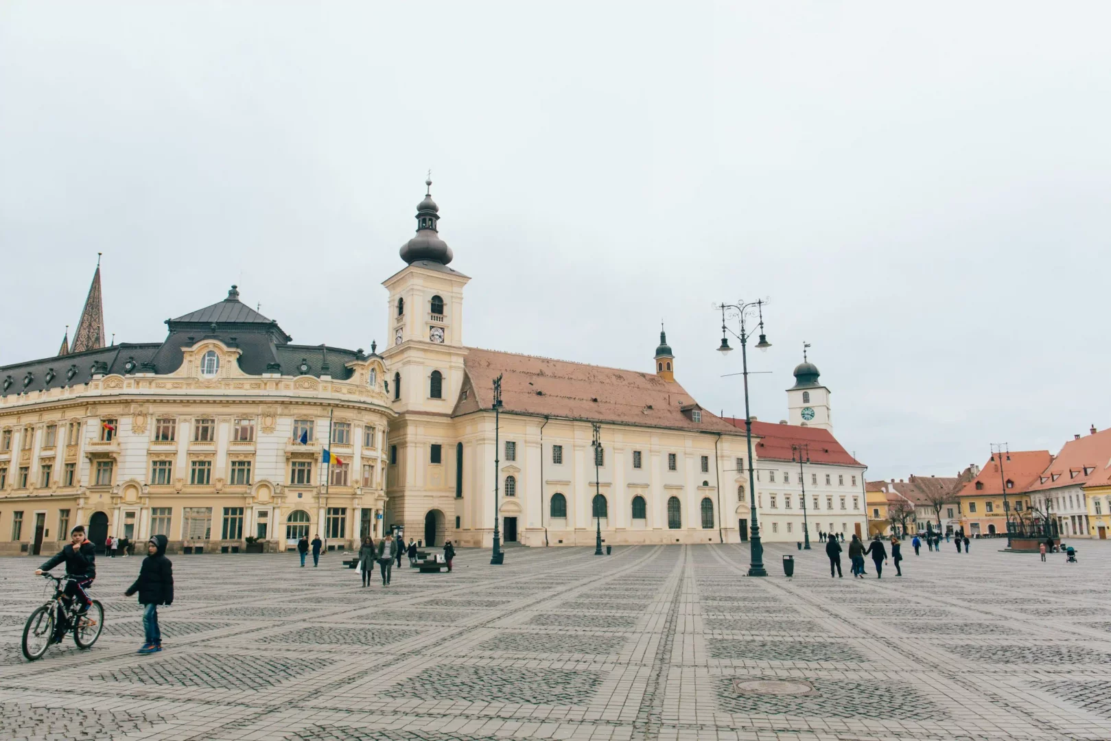 Sibiu'nun taş döşeli Büyük Meydanı'nda, sol tarafta süslü cepheli ve bayraklı tarihi bir bina, sağ tarafta ise soğan kubbeli çan kulesi olan görkemli bir kilise yer alırken, meydanda bisiklet süren ve yürüyen çok sayıda insan, gri gökyüzünün altında canlı bir şehir atmosferi oluşturuyor.