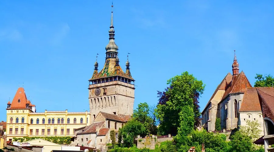 Sighișoara Orta Çağ Kalesi'nin merkezinde yer alan, renkli kiremitli çatısıyla dikkat çeken saat kulesi, yemyeşil ağaçlar ve diğer tarihi binalarla çevrili, açık mavi gökyüzü altında büyüleyici bir görünüm sunuyor.