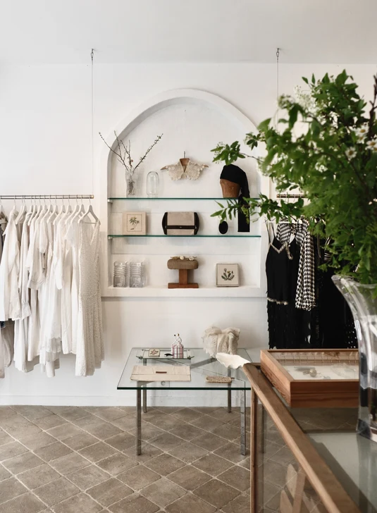 Beyaz duvarları ve kemerli niş raflarıyla dikkat çeken minimalist bir butik iç mekanında, sol tarafta zarif beyaz giysiler, sağda ise siyah ve desenli kıyafetler sergilenirken, ahşap detaylı şık cam vitrinler ve ortadaki cam masa üzerinde dekoratif aksesuarlar bulunuyor, zeminde diagonal döşenmiş taş karolar ve sağ ön planda bol yapraklı bir yeşil bitki ile sade ve modern bir mağaza atmosferi yansıtılıyor.