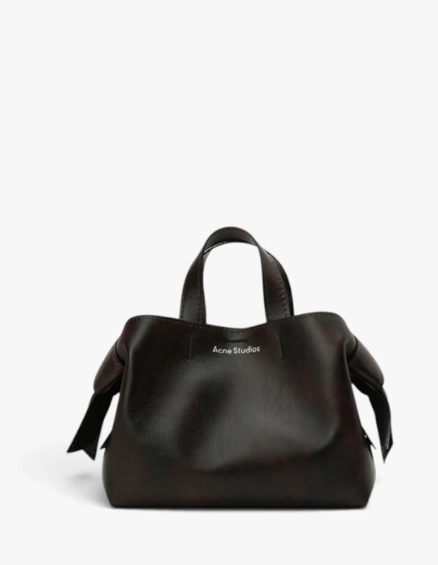 Koyu kahverengi tonlarında, yan kısımlarında karakteristik bağlamalı düğüm detaylarına sahip, iki kısa tutma saplı ve ön yüzünde Acne Studios markası belirgin olan bu şık el çantası, sonbahar-kış moda trendlerinin öne çıkan parçalarından biri olarak sergileniyor.