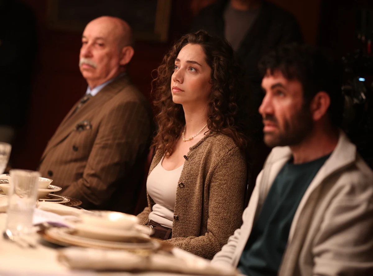 Soy filmi'nden bir karede Erdal Küçükkömürcü kahverengi çizgili takım elbisesiyle, Özge Özacar krem bluzu ve kahverengi hırkasıyla düşünceli bir ifadeyle, Mehmet Yılmaz Ak ise açık renk ceketle beyaz örtülü şık bir yemek masasında yan yana oturuyorlar.