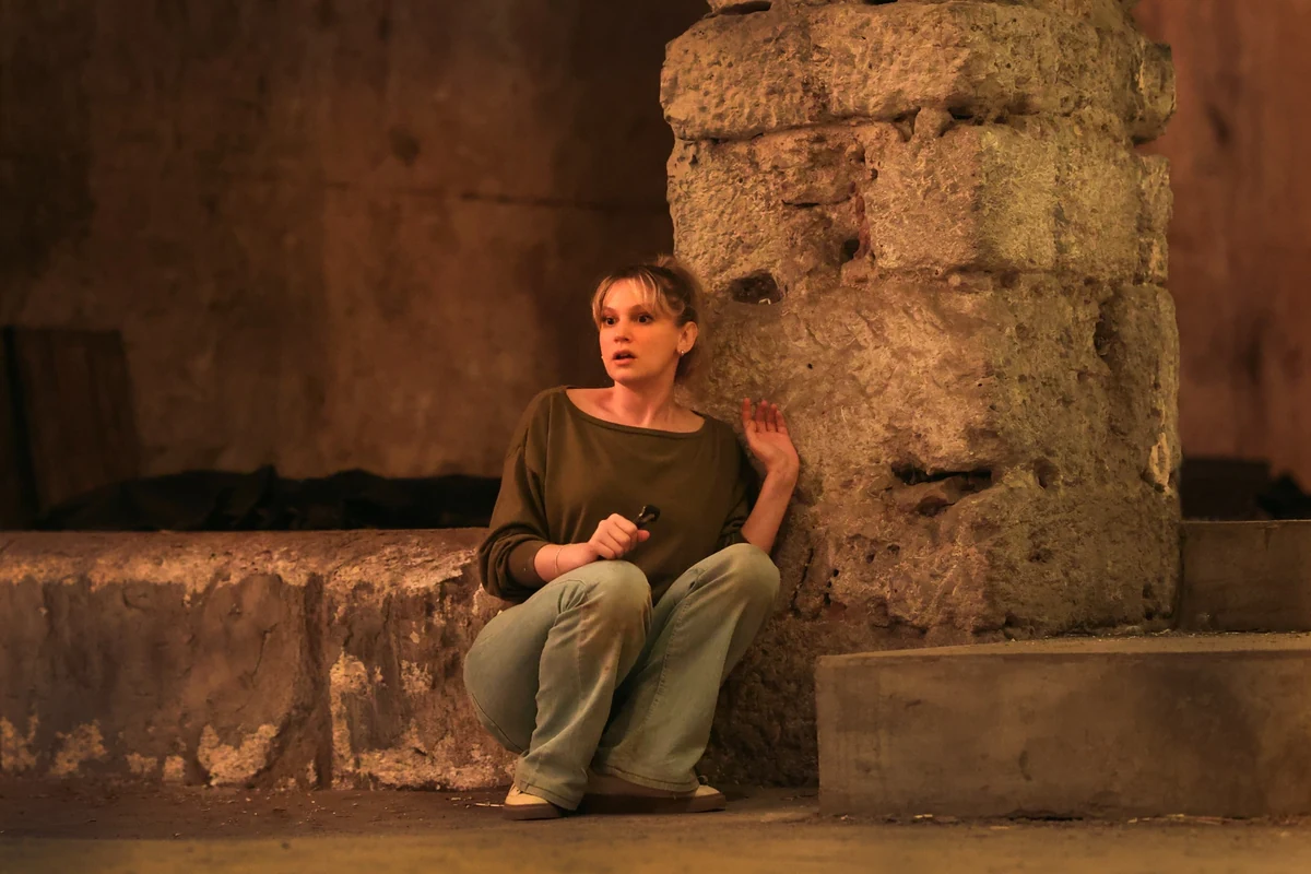 Farah Zeynep Abdullah, soy filminden bir sahnede, karanlık ve taştan bir mekanda tedirginlikle çömelmiş, ağzı hafif açık ve gözleri irice açılmış bir şekilde sağ elinde küçük bir fener tutarken sol elini kalın bir duvara yaslamış.