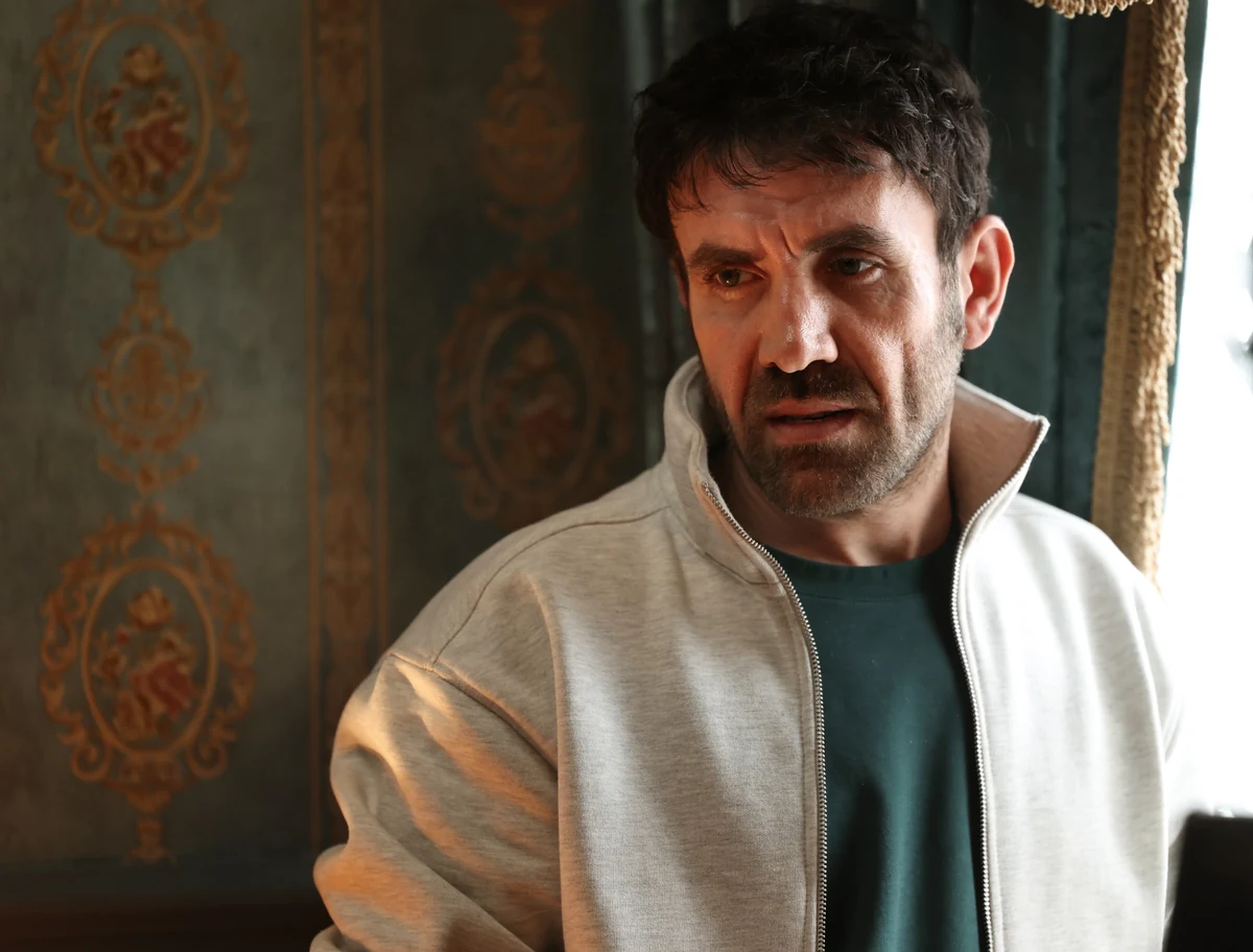 Mehmet Yılmaz Ak, Soy filminden bir karede koyu yeşil arka planda altın varaklı motiflerle bezeli duvara karşı, gri fermuarlı ceketi ve koyu yeşil tişörtüyle ciddi ve düşünceli bir ifadeyle bakıyor.