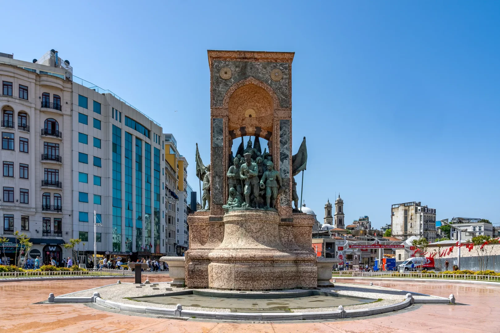 Taksim Meydanı'nın ortasında yer alan, etrafı modern ve eski binalar ile yürüyen insanlarla çevrili, açık mavi gökyüzü altındaki Taksim Cumhuriyet Anıtı, Beyoğlu kültür rotasının kamusal hafızaya açılan ilk tarihsel eşiği olarak dikkat çekiyor.