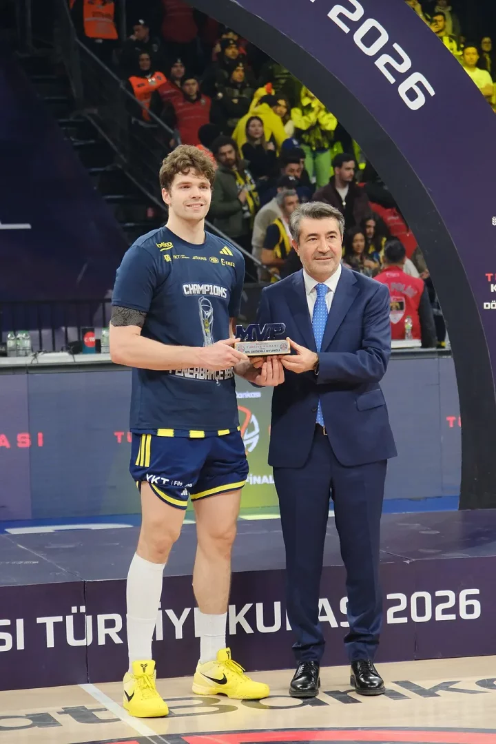 Tarık Biberoviç, koyu lacivert basketbol forması ve sarı spor ayakkabılarıyla takım elbiseli bir beyefendiyle birlikte podyumda durmuş, ikisi de gülümsüyor ve Türkiye Kupası MVP ödülünü tutuyor, arkalarında tribünler ve 2026 yazan bir kemer var.