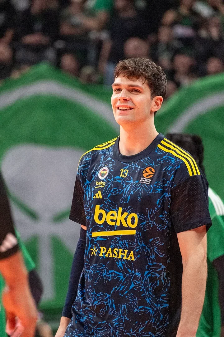 Fenerbahçe'nin 13 numaralı formasını taşıyan Tarık Biberovic, mavi ve sarı detaylara sahip forması, Beko sponsorluğu ve kulüp logosuyla parkede içten bir gülümseme sergilerken, arkasındaki flu tribünler ve yeşil bayraklar maç ortamını gözler önüne seriyor, genç oyuncunun Fenerbahçe kariyerindeki neşeli anlardan birini ölümsüzleştiriyor.