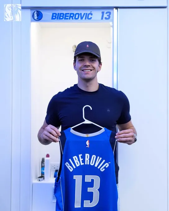Tarık Biberović, üzerinde kendi adı BIBEROVIC ve 13 numarası yazılı, mavi bir Dallas Mavericks NBA formasını askıyla birlikte tutarak soyunma odasında genişçe gülümsüyor; arkasındaki dolabın üzerinde de Dallas Mavericks logosu ve aynı ad ile numara görünüyor.