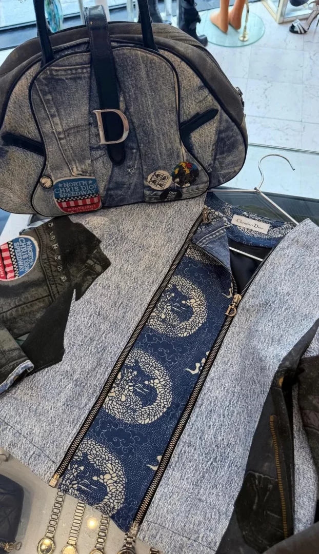 Açık gri tonlarında kot görünümlü kumaştan yapılmış, üzerinde gümüş renkli büyük bir D tokası ve NORTH CHRIS DIOR SPEEDWAY gibi çeşitli dikkat çekici yamaların bulunduğu bir el çantası ile, iç kısmında geleneksel Japon ejderha motifleriyle süslenmiş koyu mavi bir astarı ortaya çıkaran diagonal fermuarlı ceketin modern ve cesur tasarımı, güçlü bir stil ifadesi sunuyor.