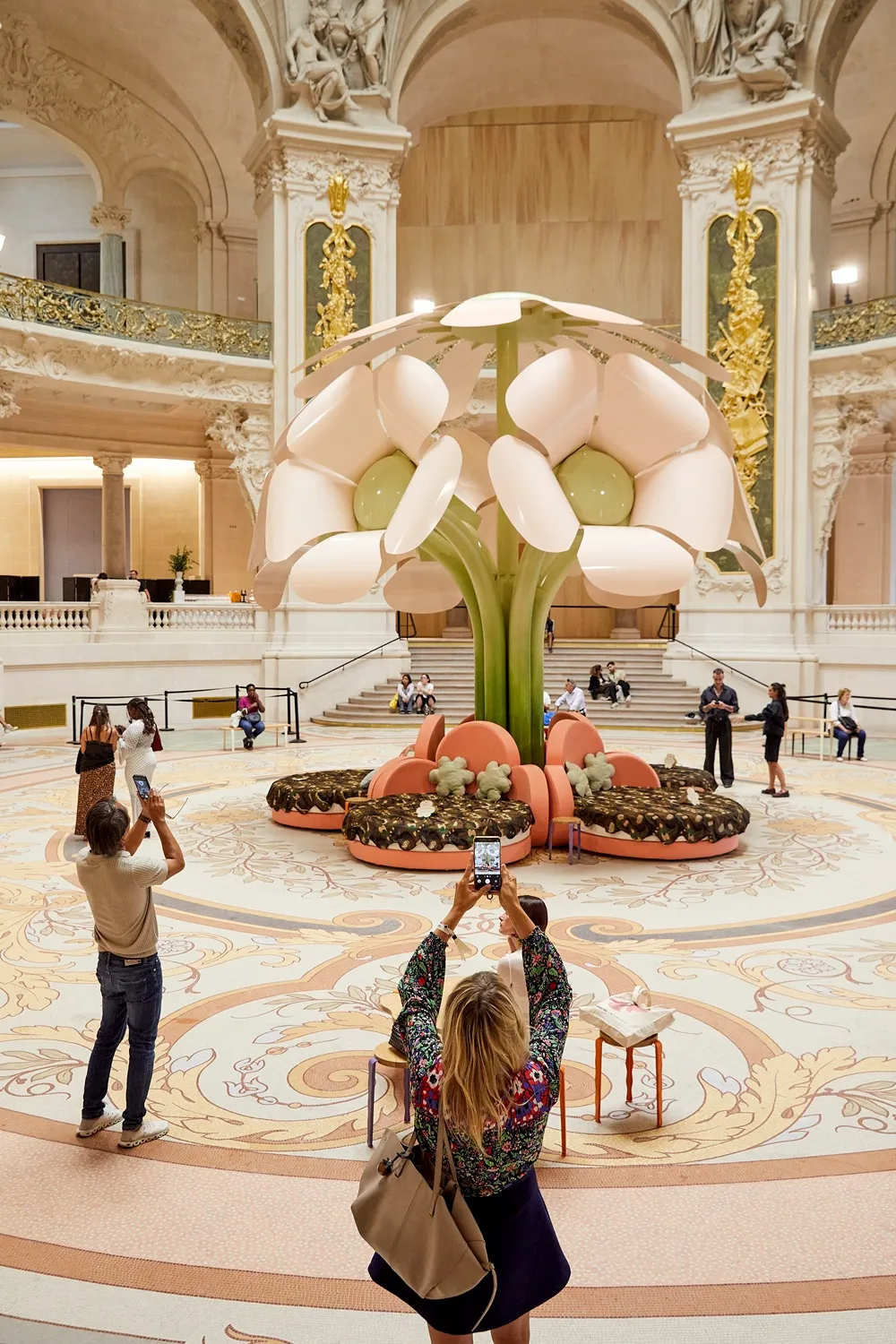 Geniş, yüksek tavanlı ve altın işlemeli detaylara sahip gösterişli bir salonda, ortada Uchronia'nın Paris Design Week için tasarladığı, açık pembe yaprakları ve yeşil gövdesi olan devasa bir çiçek formunda enstalasyon duruyor, etrafında desenli zeminde fotoğraf çeken ve dinlenen ziyaretçilerle birlikte çağdaş bir tasarım sergisini yansıtıyor.