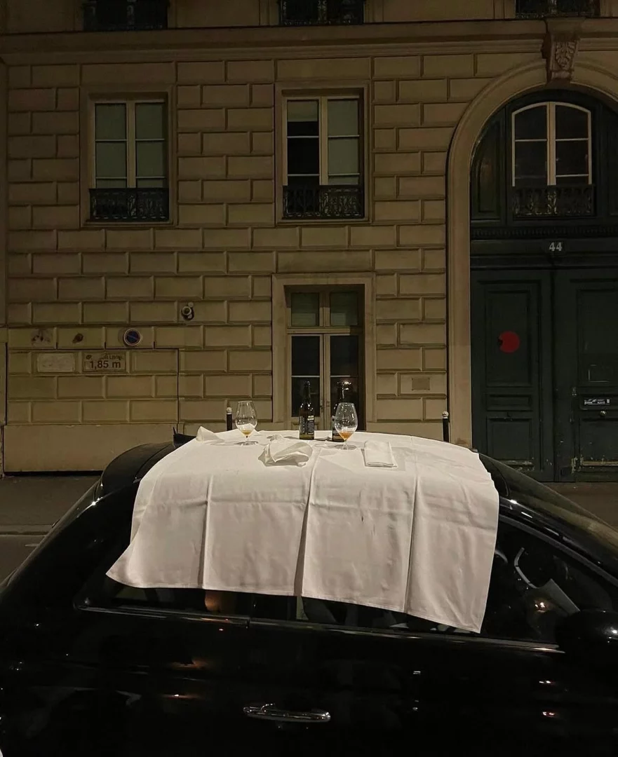 Gece ışıklandırılmış taş duvarlı bir binanın önünde parkedilmiş siyah bir arabanın çatısına serilmiş beyaz masa örtüsünün üzerinde, içinde içecek bulunan iki kadeh, iki koyu renk şişe ve katlanmış beyaz peçetelerle özgün bir açık hava yemek düzeni dikkat çekiyor.