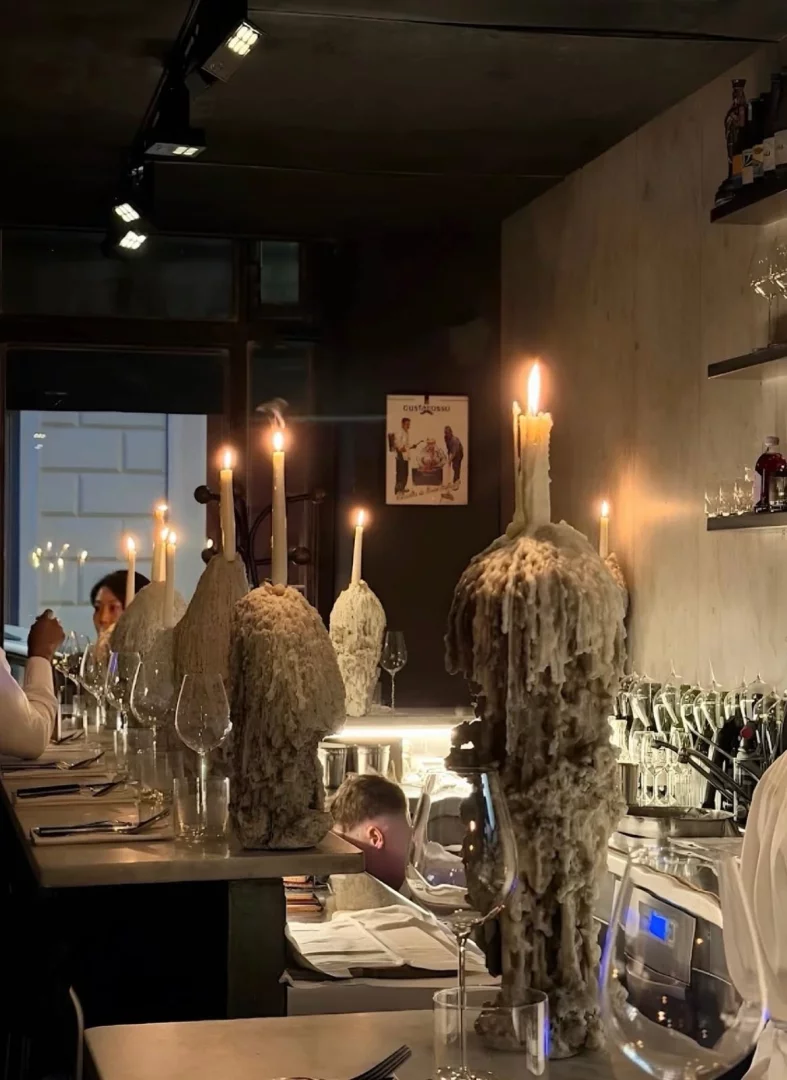 Loş ışıklı bir restoranda, uzun bir masanın üzerinde erimiş balmumundan devasa şekiller almış, ışık saçan mumlarla aydınlatılmış, şarap kadehleri ve çatal bıçak takımlarıyla dolu samimi bir yemek ortamı; arka planda hafifçe görünen müşteriler ve bir bar görevlisi, sıcak ve davetkar bir atmosfer sunuyor.