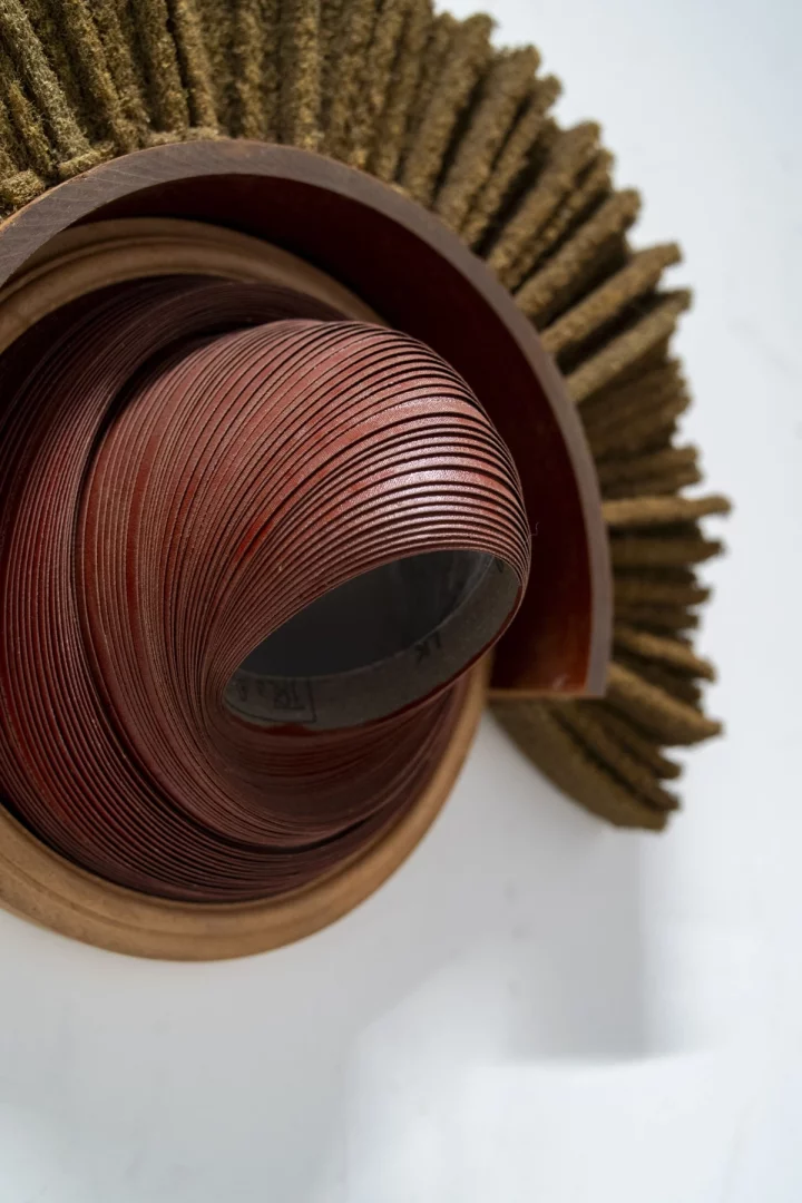 Yakın çekim bir karışık teknik sanat eserinde, merkezi kısmı saran, yoğun katmanlı koyu kızıl kahve şeritlerden oluşan sıkıca kıvrılmış bir spiral, açık renkli ahşap görünümlü bir halka ile çevrelenmiş ve sağ üst köşeden yayılan zeytin yeşili fırçamsı lifli dokularla beyaz bir arka plan üzerinde modern ve organik bir düzenleme sergileniyor.