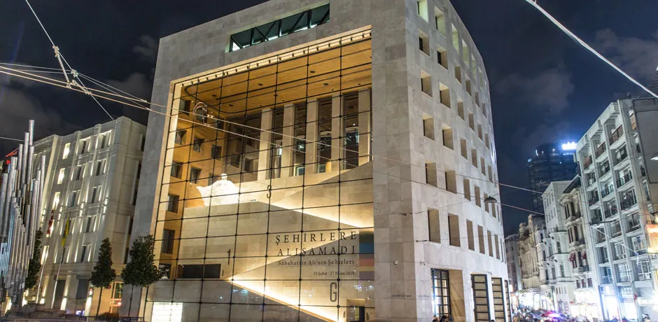 Yapı Kredi Kültür Sanat binası, gece aydınlatmalarıyla parlarken, cam cephesinde Sabahattin Ali'nin Şehirlere Alışamadı sergisine ait büyük yazılar ve heykeller görünüyor, çevresindeki şehir dokusuyla modern mimariyi birleştiriyor.