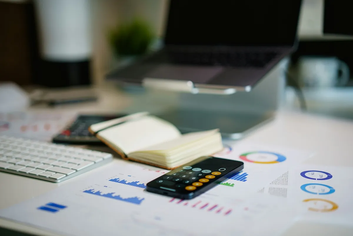 Masa üzerinde, farklı yatırım seçeneklerini gösteren detaylı grafiklerin basılı olduğu finansal raporlar, hesap makinesi uygulaması açık bir akıllı telefon ve not defteri, yatırım fonu seçimi için kapsamlı bir analiz sürecini görselleştiriyor.