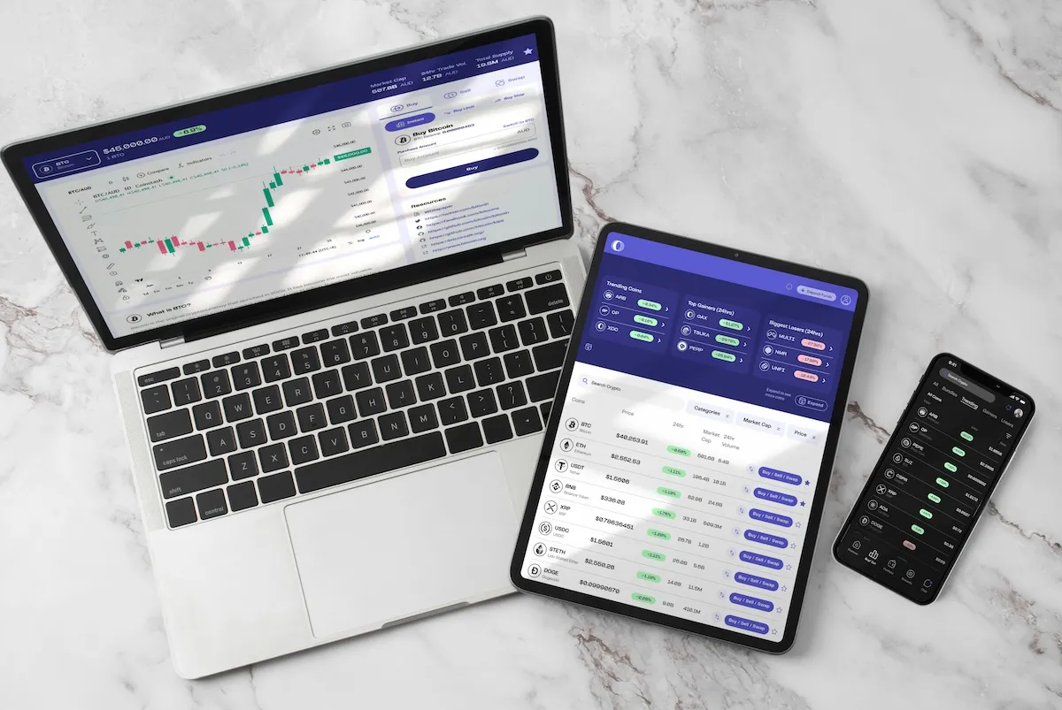 Beyaz mermer bir yüzey üzerinde duran, finansal verileri ve alım-satım ekranlarını gösteren bir dizüstü bilgisayar, tablet ve akıllı telefon, kripto para birimi piyasalarındaki yatırım fırsatlarını ve çeşitli yatırım stratejilerini dijital platformlar aracılığıyla takip etme imkanını gözler önüne seriyor.
