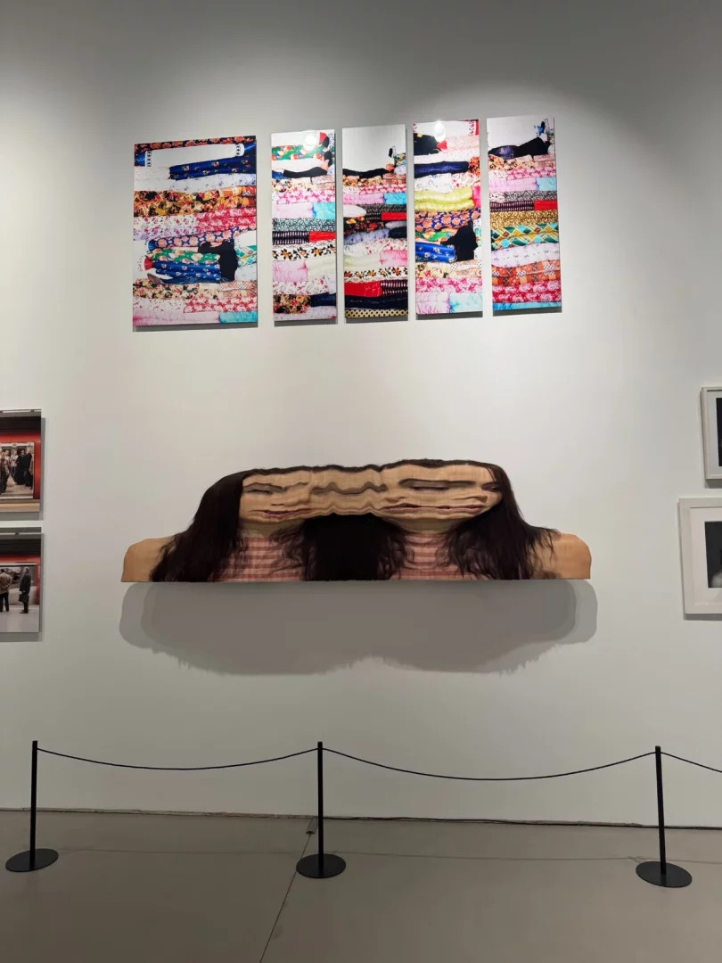 Sanat galerisi duvarında üst kısımda, renkli ve desenli kumaş yığınlarının arasında kısmen görünen insan figürlerini betimleyen dört dikey fotoğraf tablosu bulunurken, altında yatay olarak uzamış, katmanlı ve dalgalı bir formda işlenmiş, uzun koyu saçlara sahip bir kadın yüzünü gösteren çağdaş bir sanat eseri sergileniyor.